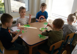 Widok na siedzące przy stoliku dzieci, które malują zieloną farbą czterolistną koniczynkę.